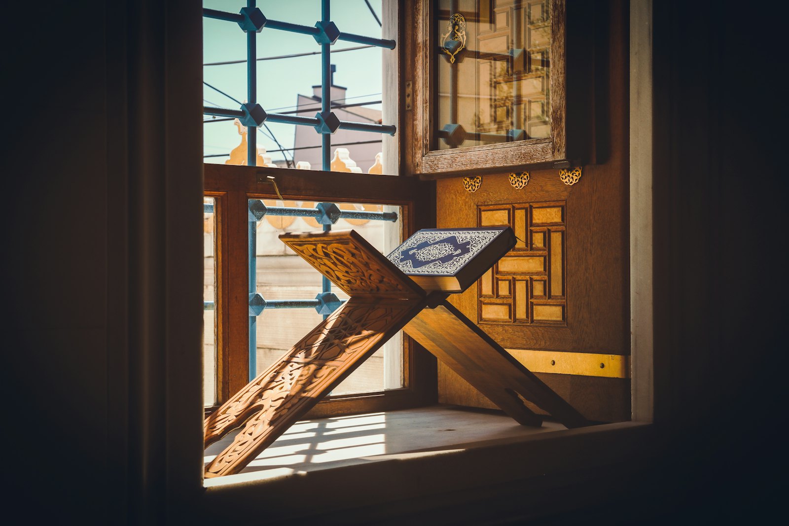 A wooden Qur'an stand holding an open book on a sunlit windowsill
